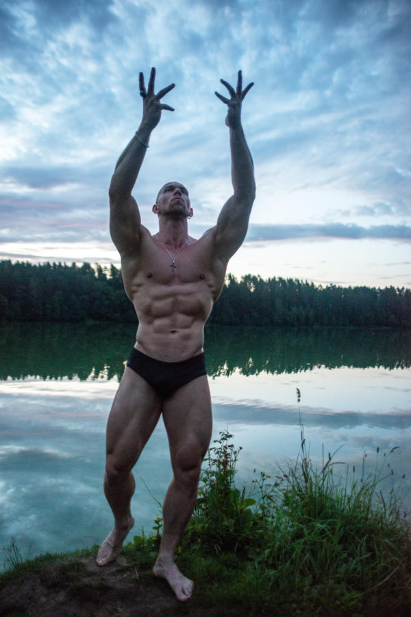 the image shows a muscular, tanned man with a welldefined sixpack and toned physique standing by a calm lake at sunset. he is posing with his arms raised overhead, showcasing his impressive muscle definition. the background features a serene lake reflecting the evening sky, which is filled with soft clouds.  the overall mood is peaceful and emphasizes the man's physical fitness and the beauty of the natural setting.  he is wearing darkcolored swim briefs.