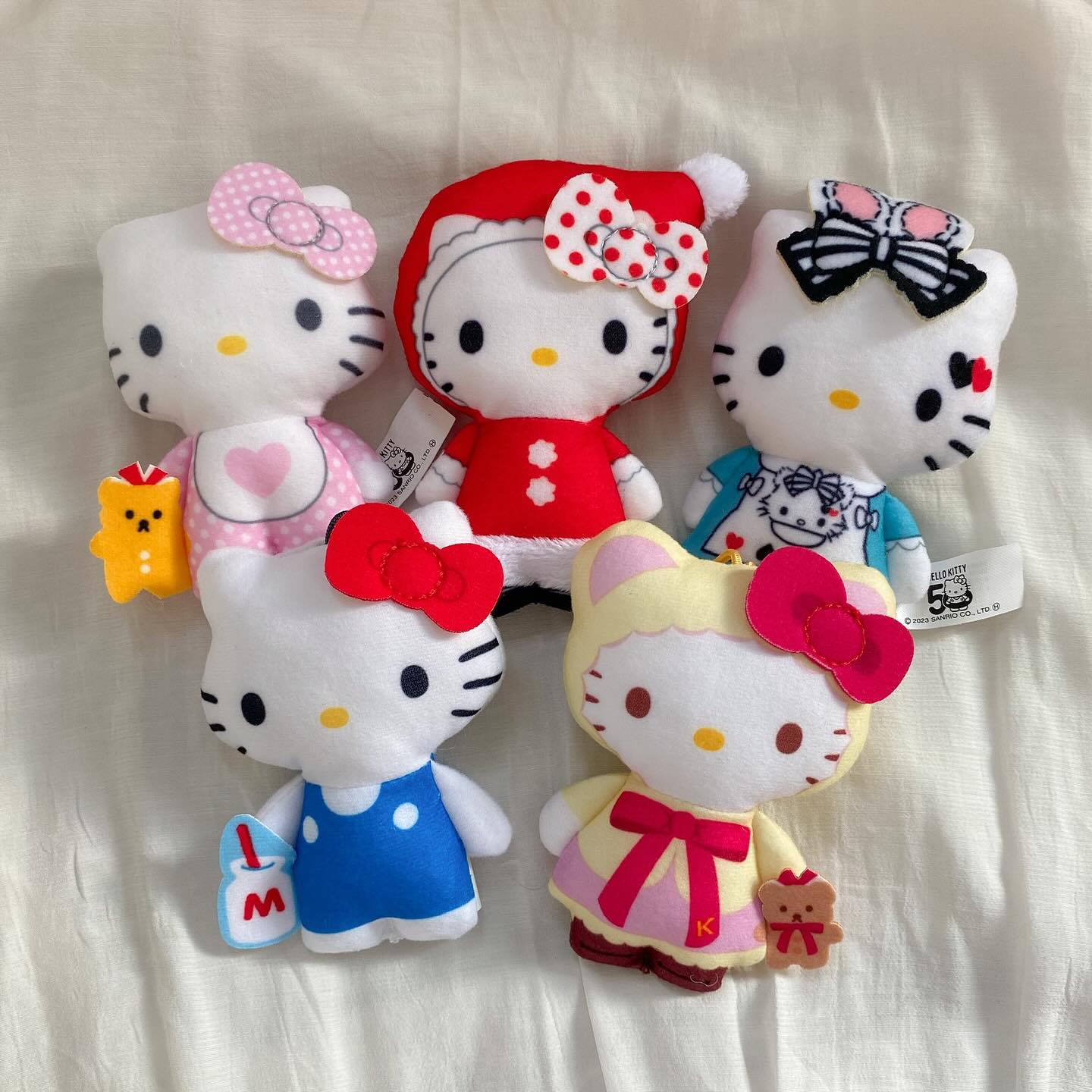 the image shows five hello kitty plush toys. the toys are arranged in a row on a white sheet. the hello kitty in the top left has a pink bow with a white circle pattern and a pink polka dot bib. she is also holding a gingerbread man. the hello kitty in the top middle is wearing a red santa suit with a white trim and a red bow with a white polka dot pattern on her head. the hello kitty in the top right has a black and white striped bow on her head. the hello kitty in the bottom left is wearing a blue overall and holding a milk carton with an "m" on the label. the hello kitty in the bottom right is wearing a yellow dress with a pink bow and holding a brown teddy bear.