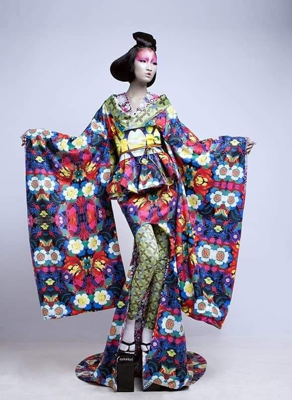 a mannequin is displayed in a vividly patterned kimono with wide sleeves, floral designs, and a peplum detail at the waist. the mannequin wears green patterned tights and high platform sandals. it has a traditional japanese hairstyle with intricate makeup. the background is a plain light gray.