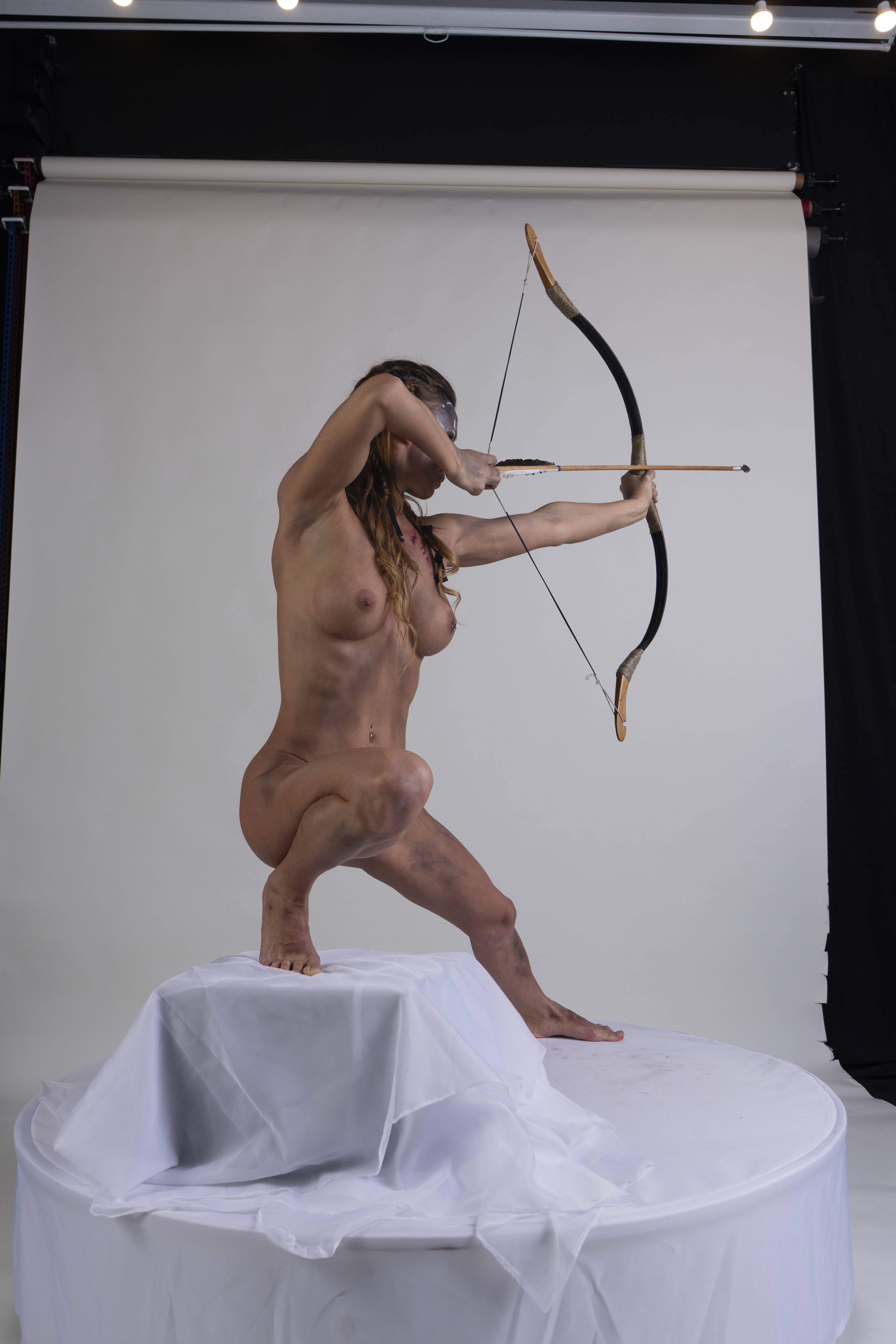 a nude woman crouches on a white platform in a studio setting, aiming a bow and arrow. she is wearing a blindfold and has a muscular physique. the background is a plain white backdrop, with some black cloth visible on the right side. the lighting is bright and even, highlighting the woman's form and the bow and arrow. she appears to be in a focused, determined stance.