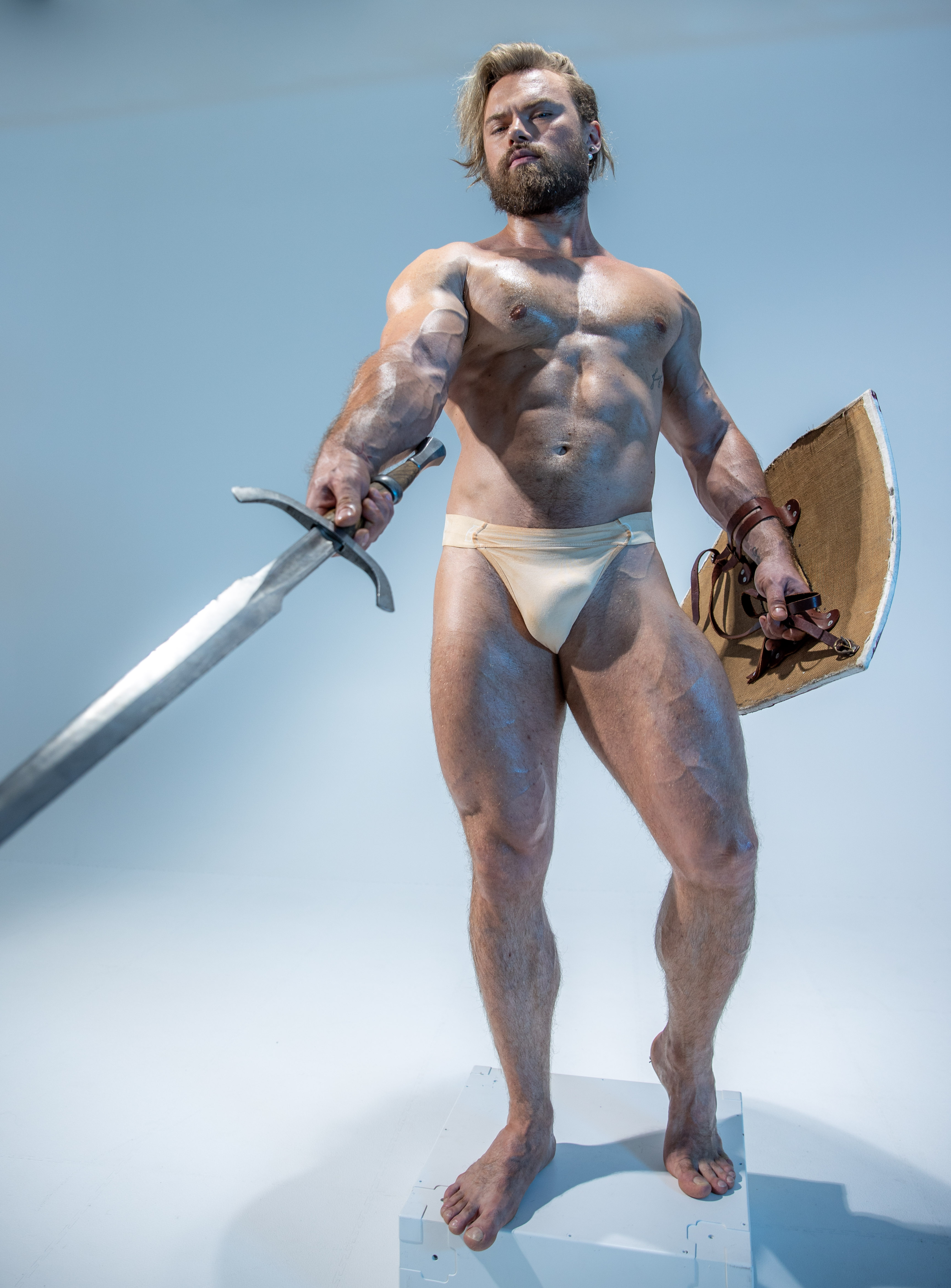 a muscular man with long blond hair and beard poses for a studio shot. he is holding a sword and shield, and is wearing a loincloth. the background is white.