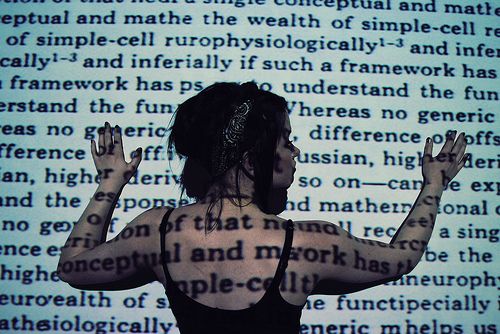 a woman with dark hair and wearing a black tank top stands in front of a wall with text projected on to it. the text covers her body and creates an abstract and intriguing image. the woman's posture and expression are mysterious, as if she is lost in thought. the overall image is artistic and captivating, leaving the viewer to wonder about the woman's story and the meaning of the text.