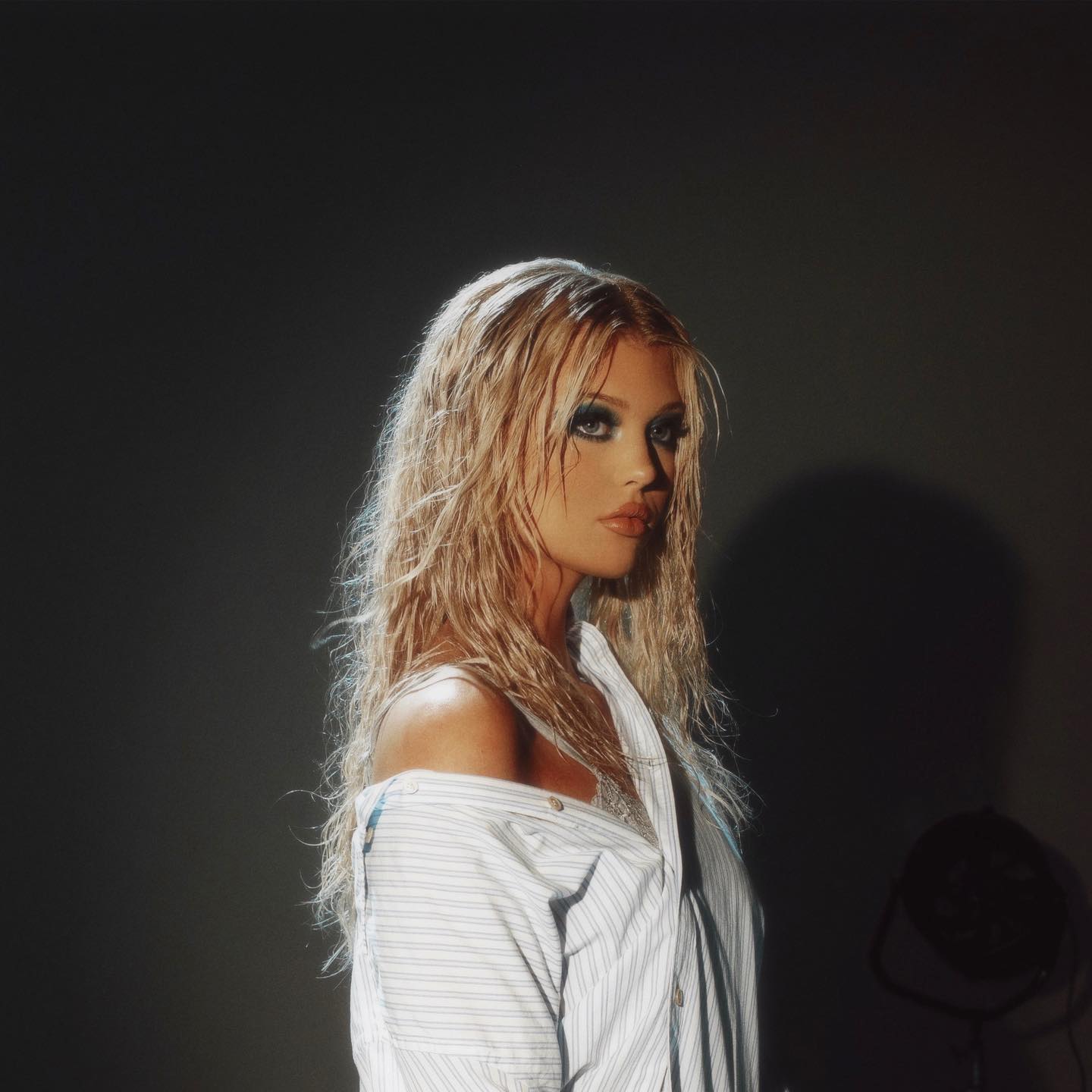 a woman with blonde, wet hair and heavy makeup is posed in front of a dark background. she is wearing a striped shirt that is partially off her shoulder. the photo appears to be a studio shot, likely a professional photoshoot.
