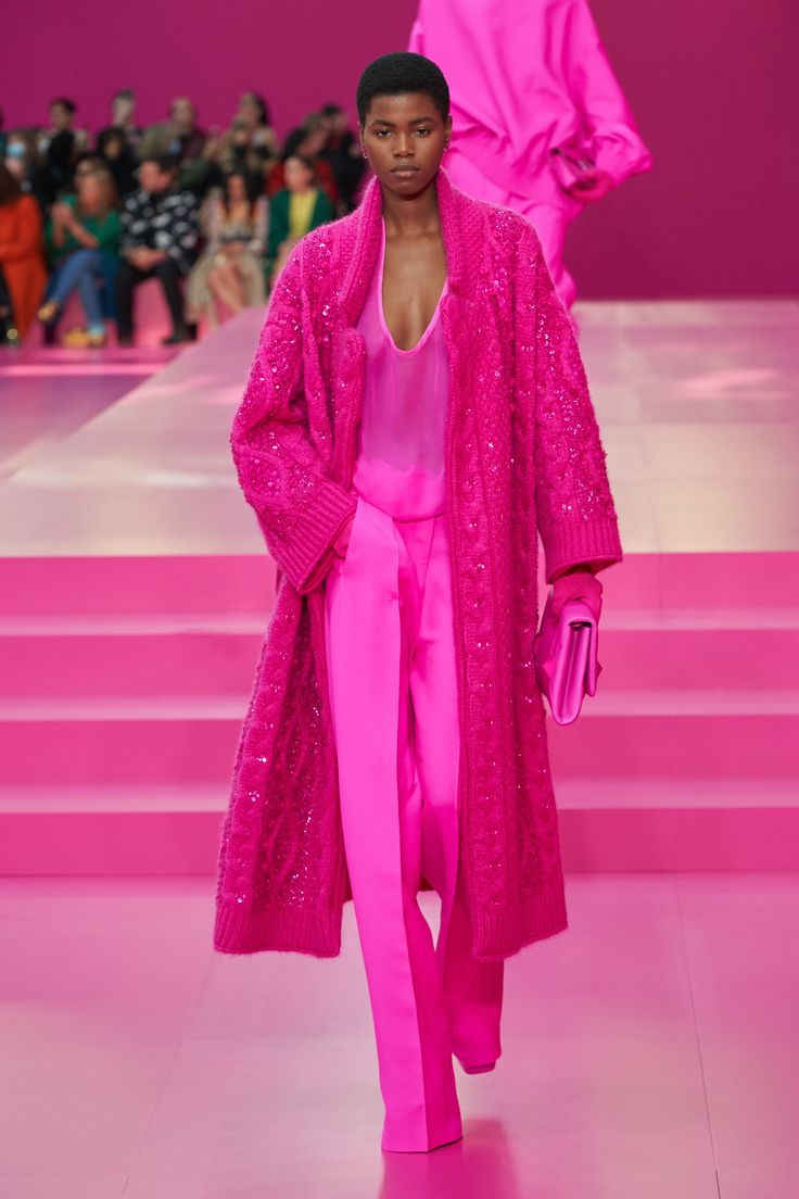 a runway shot of a black woman modeling a valentino design. she is wearing a headtotoe pink ensemble: a shimmering, cableknit cardigan over a semisheer top and matching trousers. she carries a pink handbag. the runway is a monochrome pink and a large crowd is watching.