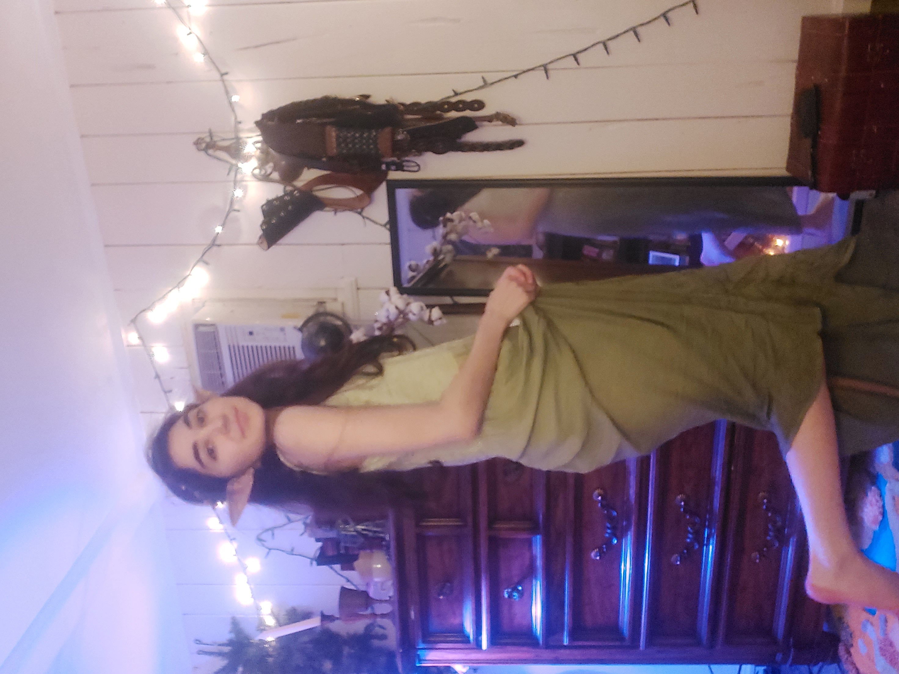a young woman dressed in an elf costume, complete with pointed elf ears, poses in her bedroom.  she wears a flowing, olive green dress and is standing in front of a wooden dresser with fairy lights strung along the wall behind her. a mirror reflects her back and a portion of the room.  the overall atmosphere is whimsical and bohemian.