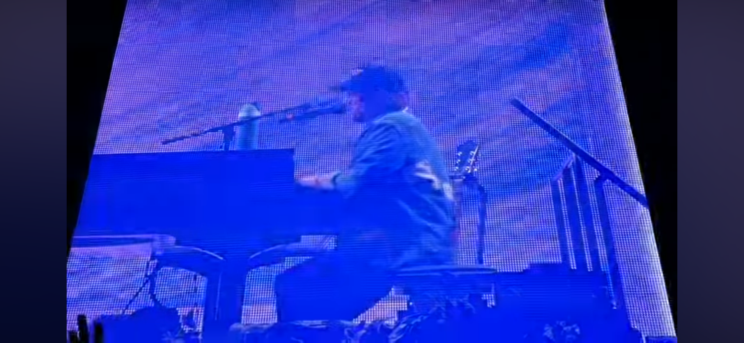 this is a blurry image of a male musician playing a keyboard on stage. the musician is wearing a hat and jacket, and there is a microphone in front of him. the stage is lit up with blue lights, and there are some shadows in the background. it looks like the musician is playing a song to an audience of people.