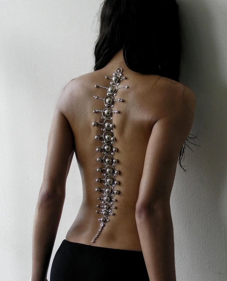 a woman is standing with her back to the camera. she has dark hair and is wearing black pants. a silver spinelike piece of jewelry is displayed on her back.