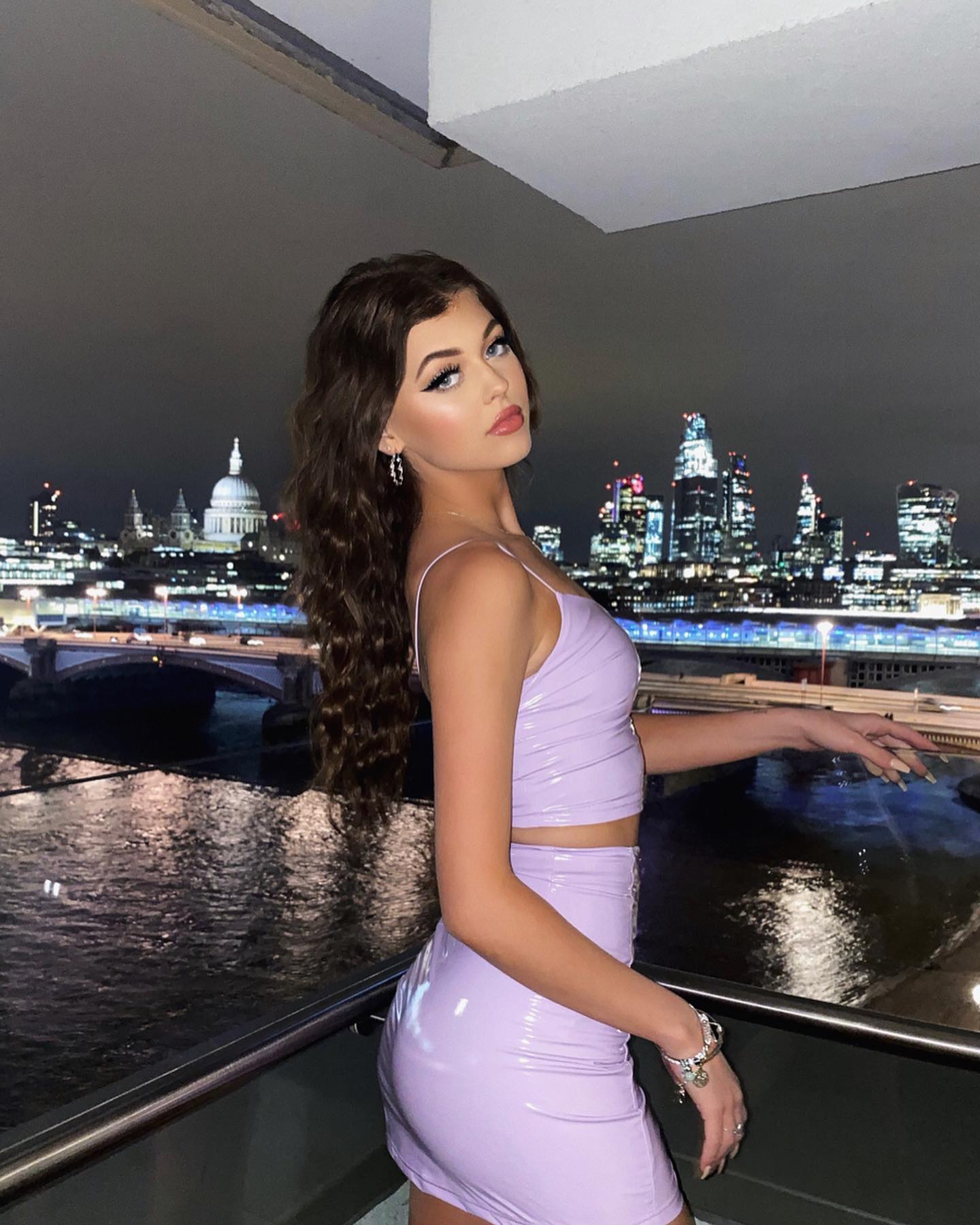 a young woman is posing on a balcony in front of a nighttime cityscape. she is wearing a shiny, light purple twopiece dress consisting of a crop top and a short skirt. she has long, curly brown hair and is wearing jewelry including a bracelet and earrings. the background shows the city of london at night, with a river and multiple bridges in the foreground and illuminated city buildings including st. paul's cathedral in the distance.