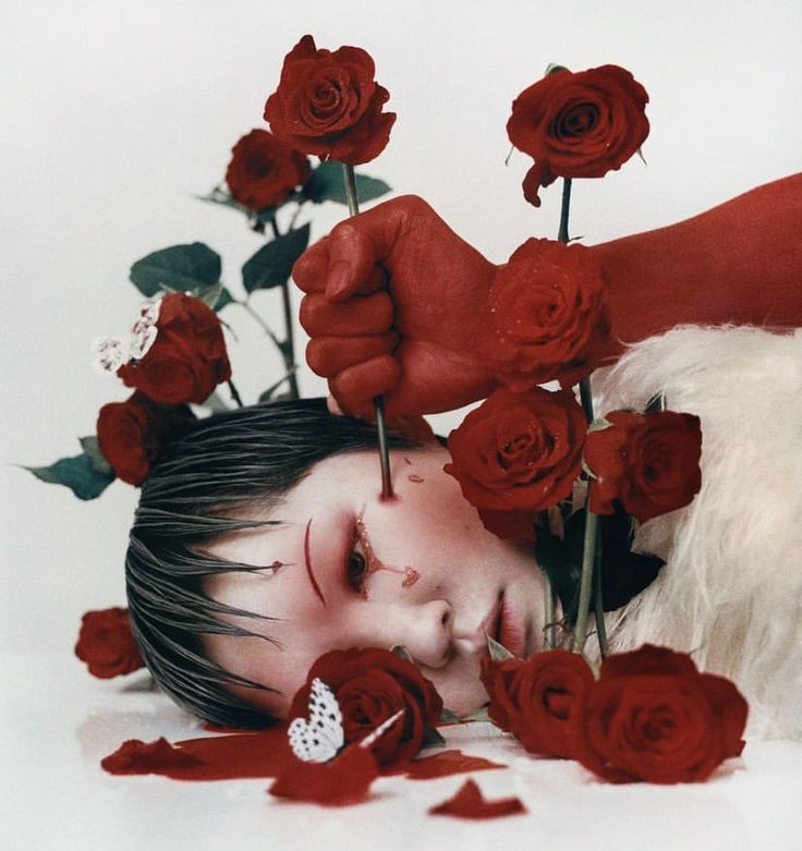the image is a dramatic and surreal closeup shot of a person's face partially obscured by red roses.  a redpainted hand holds several rose stems, some of which appear to be piercing the model's skin, with drops of red liquid suggesting blood. the model's makeup is stylized, with red accents mimicking the blood and roses. the background includes white fur and more red roses scattered around the model.  white butterflies add a contrasting element. the overall aesthetic is dark, artistic, and evokes themes of pain, beauty, and possibly mortality. the photography style is highly stylized and suggestive of a fashion or art photograph.