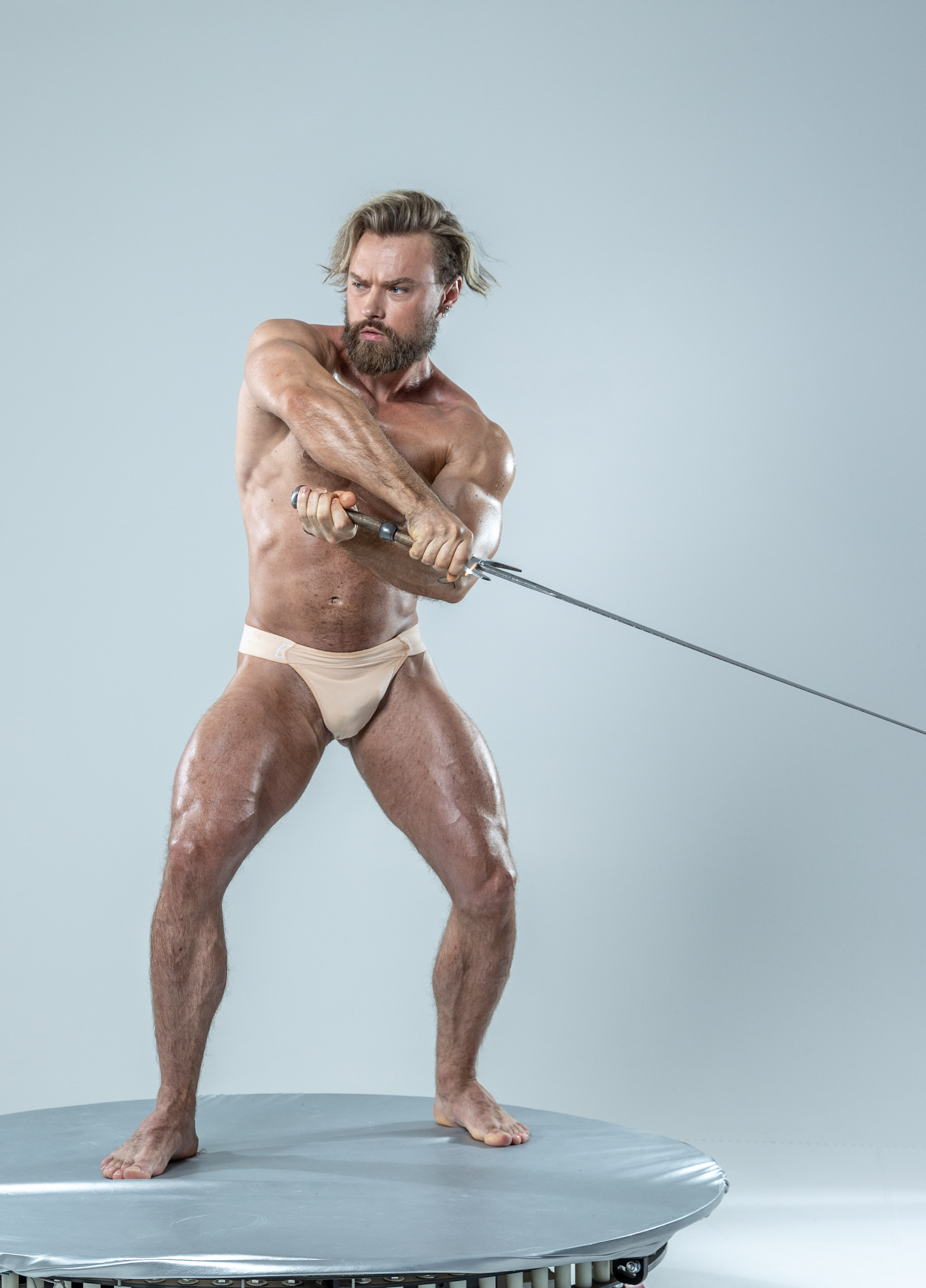 a muscular, barechested man with long blond hair and a beard stands in a fighting stance on a circular platform. he grips a sword with both hands, preparing to strike. he is lit by artificial studio lighting against a plain grey background, highlighting his physique.