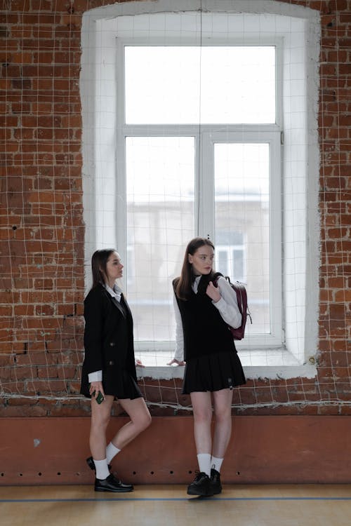 two teenage schoolgirls in uniform stand side by side against a brick wall with a large window behind them. one girl wears a black blazer and skirt with a white shirt, and carries a backpack.  the other girl wears a similar uniform but with a black vest. they appear to be casually leaning against the wall near the window. the overall setting looks like a school hallway or gymnasium.
