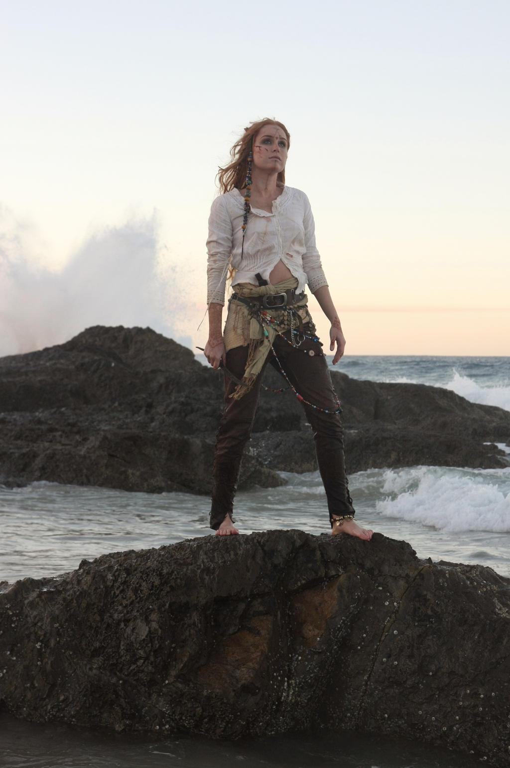 a full shot of a woman with long, braided red hair, wearing a creamcolored shirt and brown pants, stands on a dark rock near the ocean.  the waves are crashing against the rocks, and the sky is a soft, twilight color. she appears to be in a costume of a mystical or adventurous nature. the overall feel is one of fantasy or storytelling.