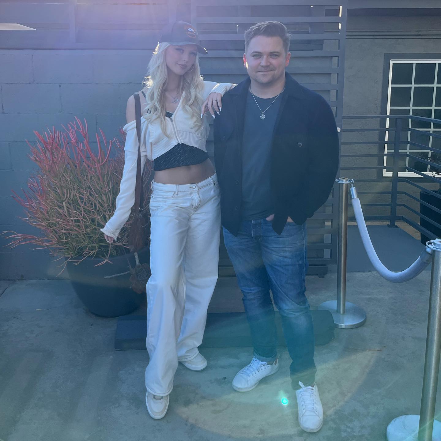 a full shot shows a woman standing next to a man outside of a building. the woman has blonde hair and is wearing a cap, a white sweater, a black tank top, white pants, white sneakers and a shoulder bag. the man has brown hair and is wearing a corduroy jacket, a dark shirt, jeans, white sneakers and a silver chain.