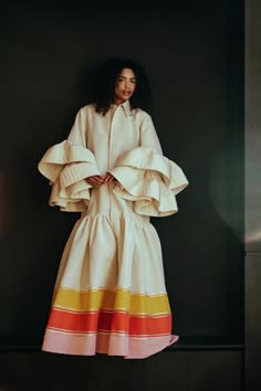 a woman is posing in a white dress with large, ruffled sleeves and stripes at the bottom. she is standing against a black background.  the woman is wearing a white dress with a white shirt collar. the dress has a lot of ruffles on the sleeves and skirt. there are three pink stripes, three yellow stripes, and two orange stripes around the bottom of the skirt.  the woman's hair is dark brown and wavy. she is looking at the camera.  the background is a black wall.