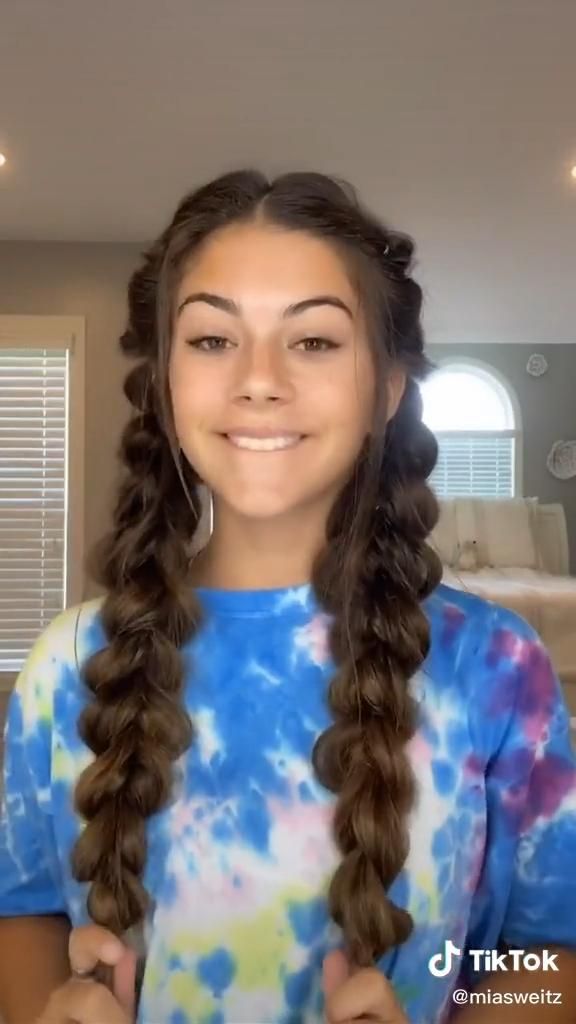 a young woman with brown hair in bubble braids smiles at the camera. she is wearing a blue and white tiedye shirt. the background is an interior setting with a window, blinds, and furniture. a tiktok logo is visible in the bottom right corner.