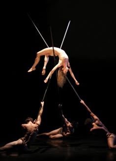 a group of dancers perform an aerial circus act. one female dancer hangs upside down, suspended from a pole. she is surrounded by other dancers who are moving around the stage. the dancers are lit by a spotlight. they appear to be athletic and graceful. the photograph captures the energy and beauty of their performance.