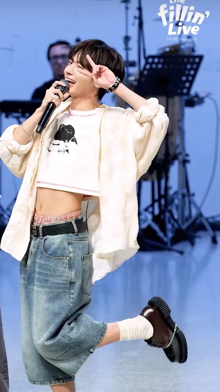 a man is singing into a microphone while giving a peace sign. he is wearing a white crop top with a black illustration, an open offwhite buttondown shirt, lightwash baggy denim shorts, brown shoes, and white socks. he has a black beaded bracelet on his left wrist and a black belt holding up his shorts. the stage setting includes musical instruments and another individual blurred in the background. the man is smiling and winking, looking directly at the camera.