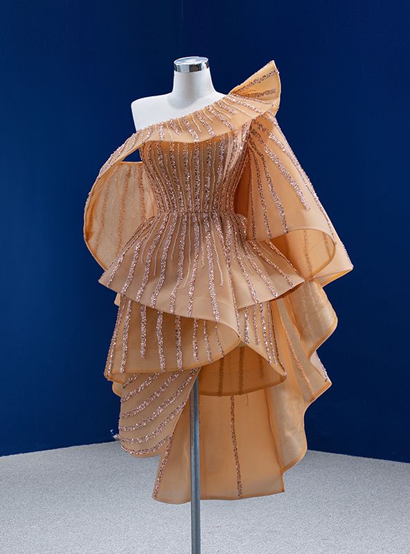 a tan and gold high low dress with ruffled detailing and a one shoulder neckline. it is displayed on a mannequin against a blue backdrop.