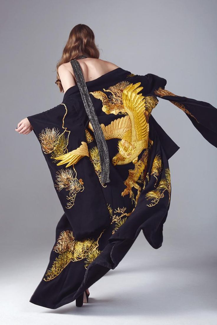 a back view of a model wearing a long, black kimono adorned with intricate gold embroidery depicting eagles and pine trees. the kimono is flowing gracefully, and the model is wearing black heels.  the setting is a simple, light grey studio background. the image emphasizes the luxurious texture and design of the kimono.