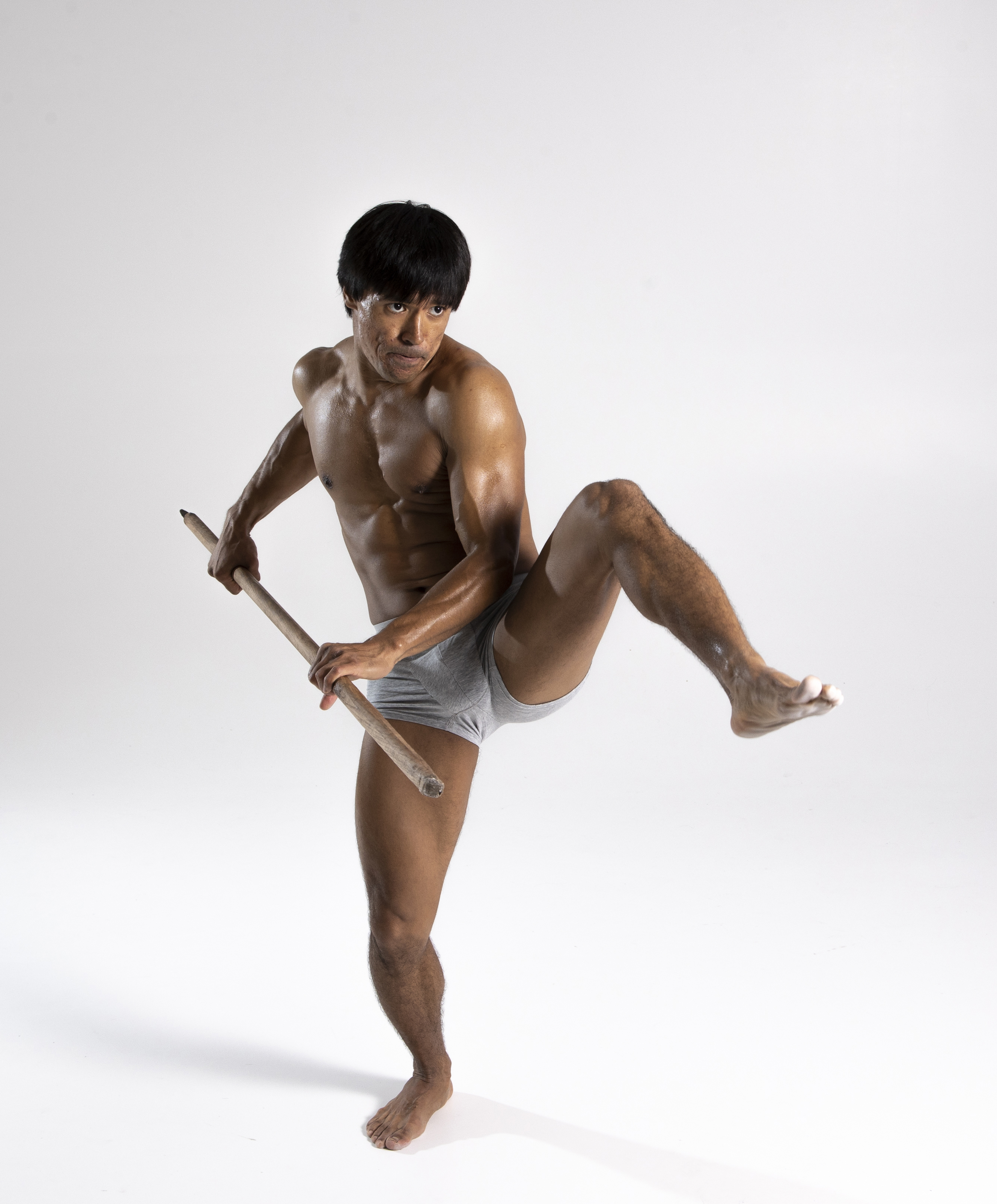 a muscular, shirtless man in grey underwear is holding a pole and standing on one leg in a martial arts pose against a light background. he is barefoot and appears to be performing a training exercise.