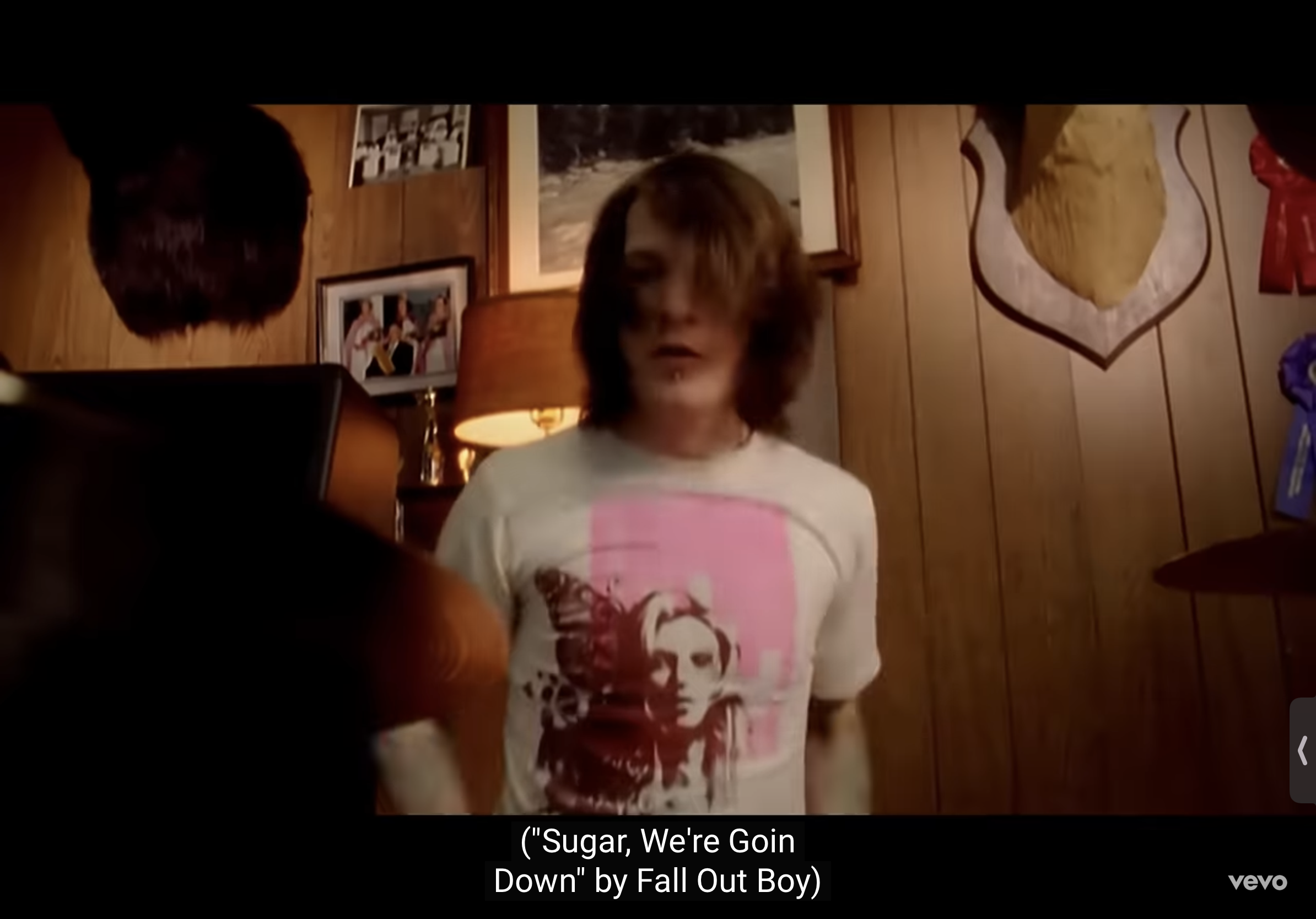 the image shows pete wentz from the band fall out boy in the music video for "sugar, we're goin down." he's wearing a white tshirt with a graphic print on it and is standing in a room with woodpaneled walls. there's a lamp with a yellow shade on the left side of the image, and a mounted deer head on the right. the lighting is dim and artificial, giving the scene a slightly gritty feel.