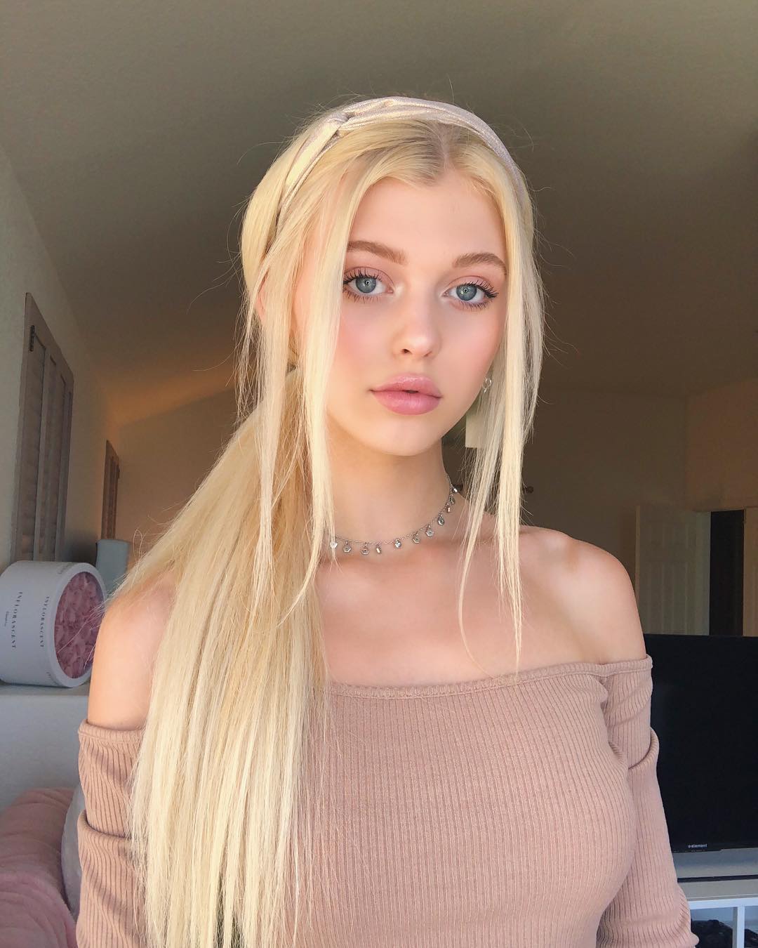 a portrait of a young woman with blonde hair and blue eyes. she is wearing a headband and a choker necklace. she is also wearing an offshoulder top. the background is neutral. the woman appears to be loren gray.