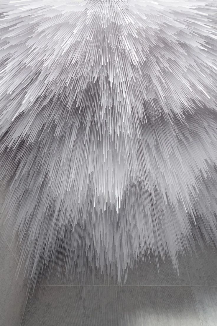 this is a closeup image of an abstract installation, most likely a ceiling, made of white lines that create a 3d pattern. the lines are arranged in a radial fashion, with their endpoints converging towards the center. the background is a light grey concretelike surface, which contrasts with the brightness of the white lines. the image is minimalist and modern in its aesthetic.