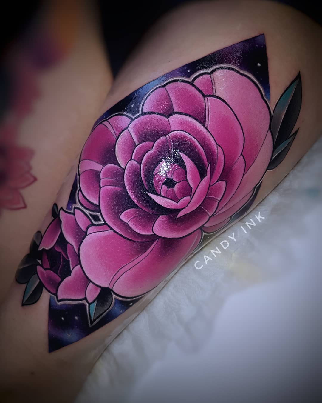 this is a closeup image of a colorful thigh tattoo of two pink roses against a galaxy background. the tattoo is done in a realistic style with bold lines and shading. the roses are the focal point of the image, and the galaxy background adds depth and dimension. the tattoo is on the outer right thigh of a person. only part of the leg is visible, and we can see the tattoo artist's table underneath. the image is lit with artificial light.
