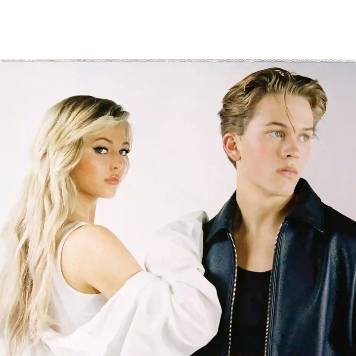 a young woman with blonde long hair and heavy eyeliner stands next to a young man with slicked back hair. she is wearing a white shirt, and he is wearing a black leather jacket. they are both in a studio shot with a white background. the woman is looking at the camera, while the man is looking off to the side.
