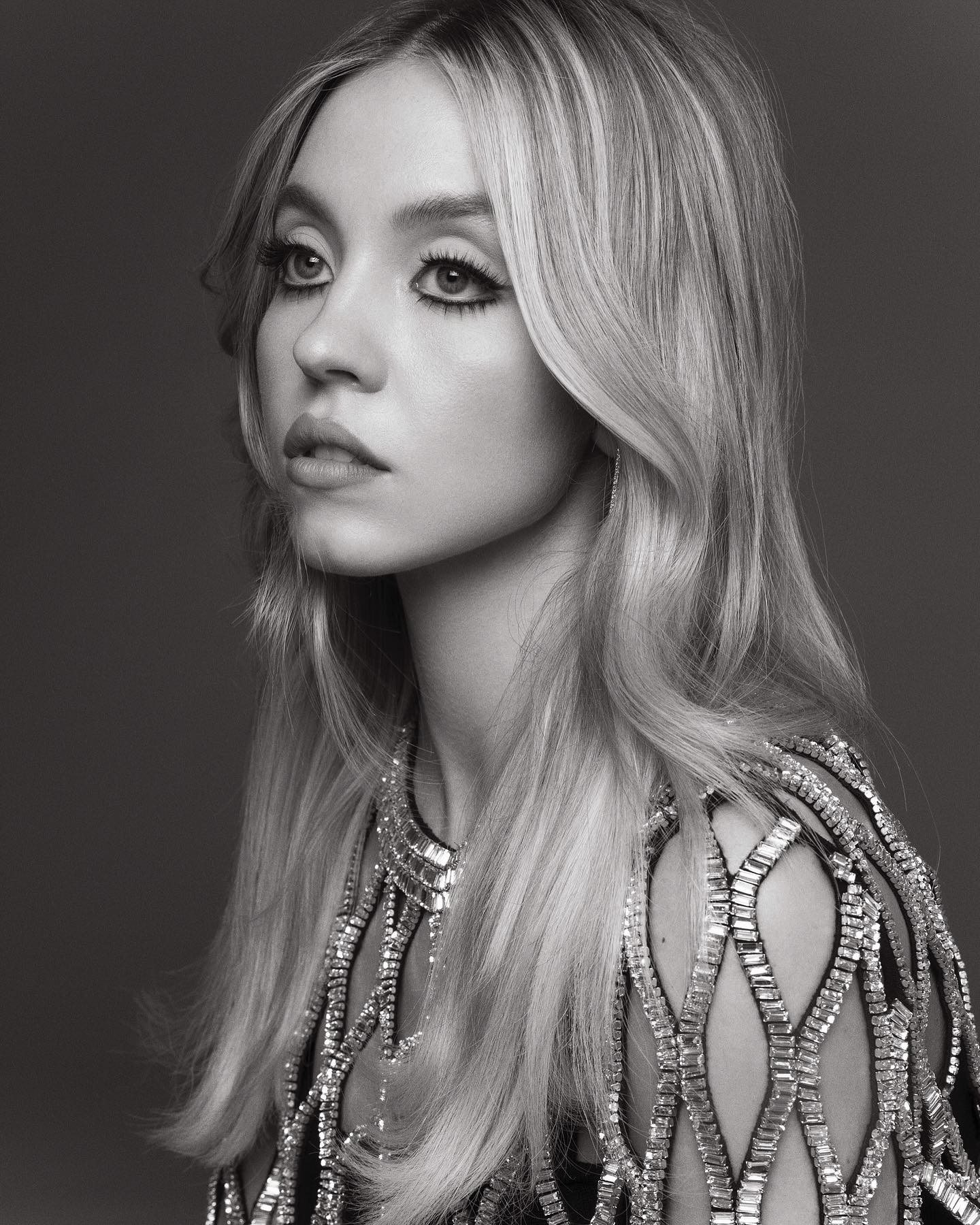 the black and white image is a closeup portrait of a young woman with long, flowing blonde hair styled in a 70sinspired fashion. her hair falls over her shoulders and partially obscures her neck. she is wearing a dark dress adorned with intricate crystal or beaded embellishments that create a latticelike pattern across the shoulders and upper bodice. her makeup features noticeable winged eyeliner.  she is looking away from the camera, creating a pensive or thoughtful expression. the background is a solid, dark gray, which helps the subject stand out. the overall mood of the photo is sophisticated and glamorous, hinting at a high fashion editorial or campaign.
