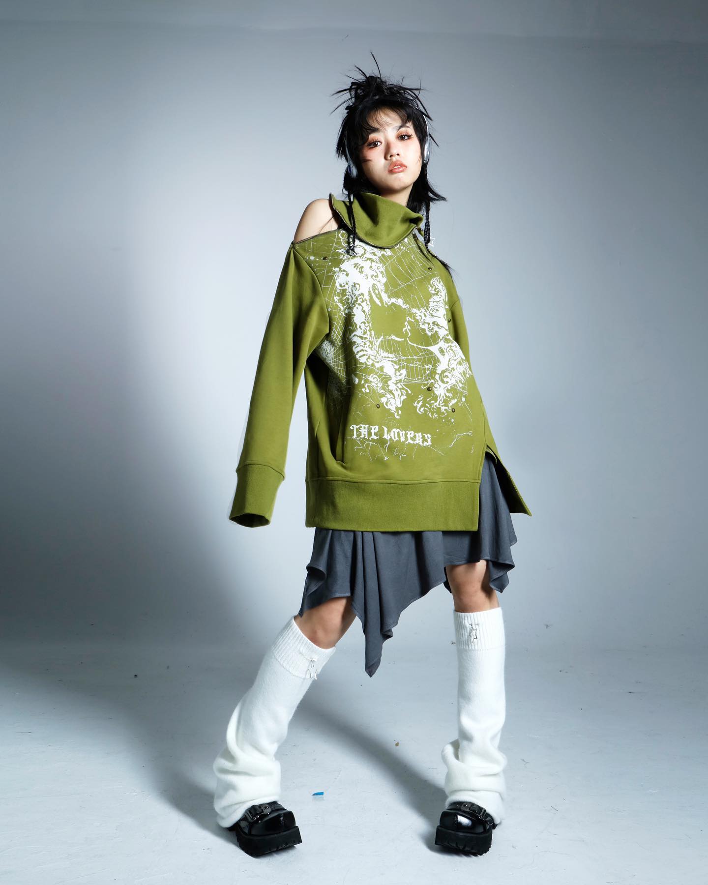 a full shot of a young woman model posing in a studio against a light grey background. she is wearing an olive green oversized sweatshirt with cutout shoulder detail and a graphic print, paired with an asymmetrical dark grey miniskirt. her styling is completed with white leg warmers and chunky black boots, and her hair is styled in a spiky updo. the overall style is edgy and contemporary.