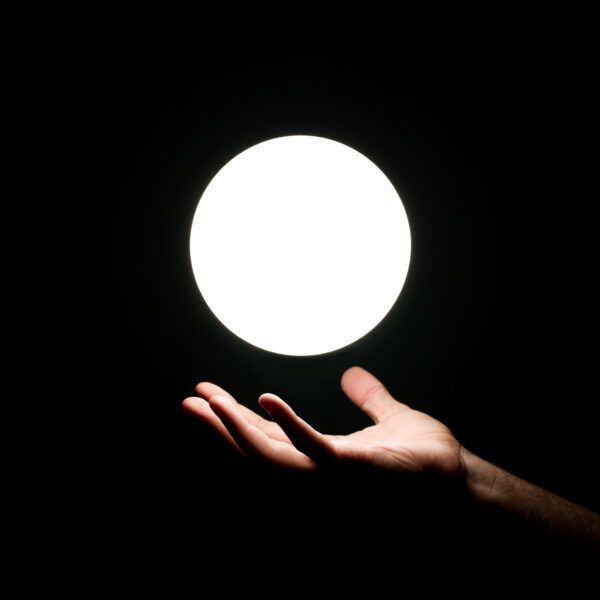 a hand is illuminated in the dark, positioned below a bright, glowing orb. the orb appears to be floating above the open palm of the hand. the background is black.