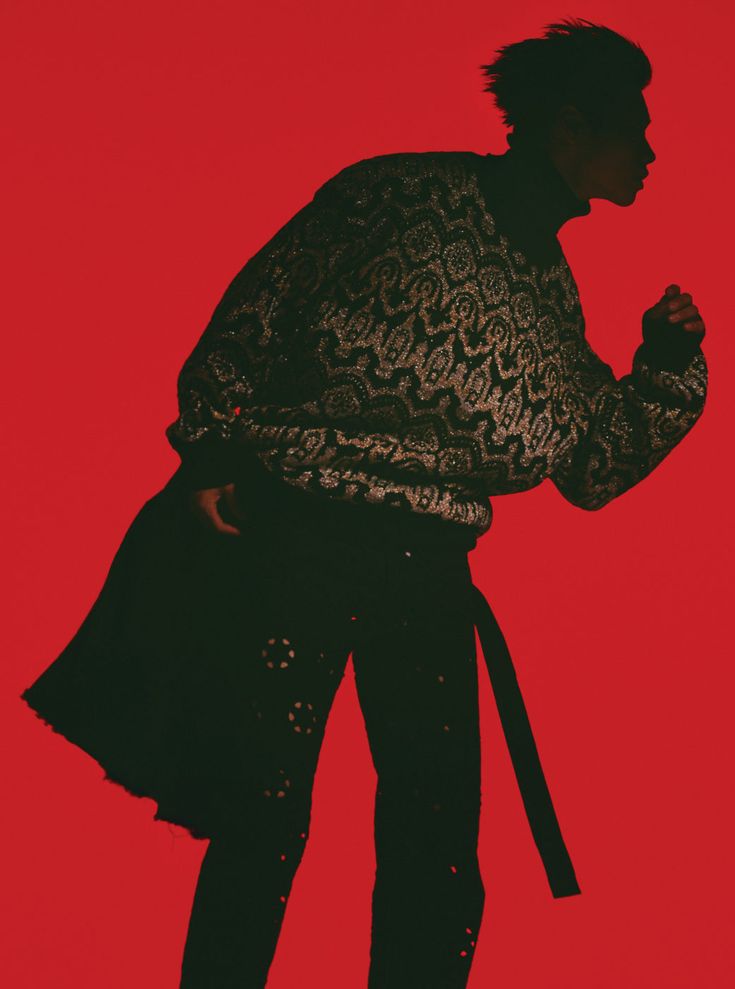 a silhouette of a person wearing a black sweater with a pattern, black jeans, and a black belt. the figure is facing away from the camera and to the right, with their right arm raised in a pose. the image has a red background.
