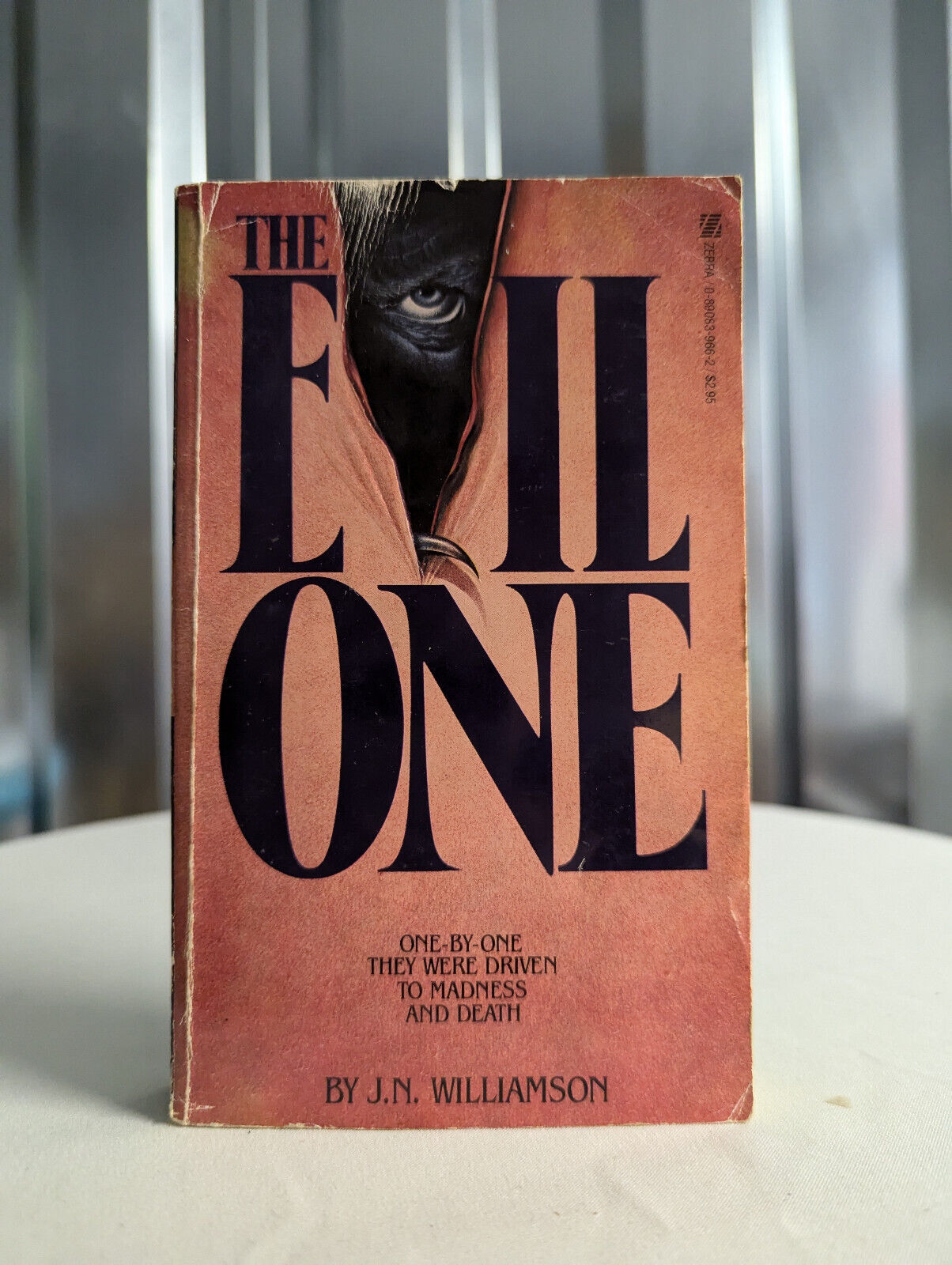 a closeup shot of a vintage paperback book titled "the evil one" by j.n. williamson. the book cover features a dark, mysterious image of an eye peering from behind a hood or cloak. the background is blurred and shows a metallic gray surface. the book is sitting on a white table. the text on the cover is clear and legible.