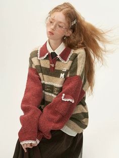 a young woman with long red hair and glasses is posing in a studio, wearing a trendy oversized striped sweater with a collar and lace detailing. the sweater is a mix of green, brown, and cream stripes with red sleeves.  she is wearing a white shirt underneath and a darkcolored skirt.  the photo is welllit and shows off the details of the clothing.