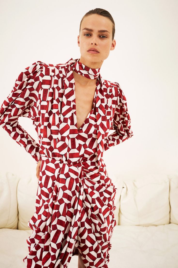 a woman is standing on a white couch, wearing a longsleeved, red and white geometric print dress. the dress has puff sleeves, a high neck, and a draped design at the waist. the dress's print is a repeating pattern of red and white geometric shapes. the model has her hair pulled back and wears minimal makeup. the background is a plain white wall.
