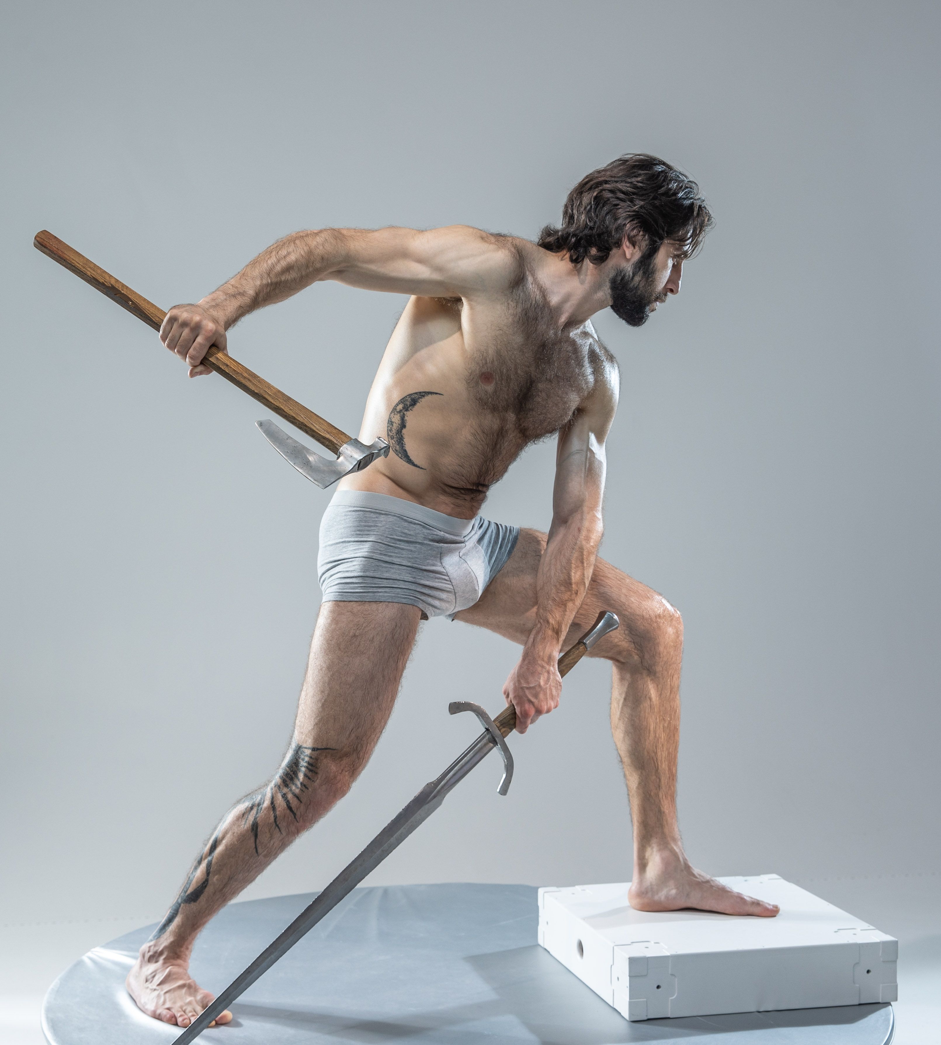 a muscular, bearded man in grey underwear poses dramatically on a white platform against a plain grey background. he holds a shorthandled axe in his right hand, raised above his head, and a sword in his left hand, pointed towards the ground. the image is brightly lit in a studio setting, likely for artistic or modeling purposes.