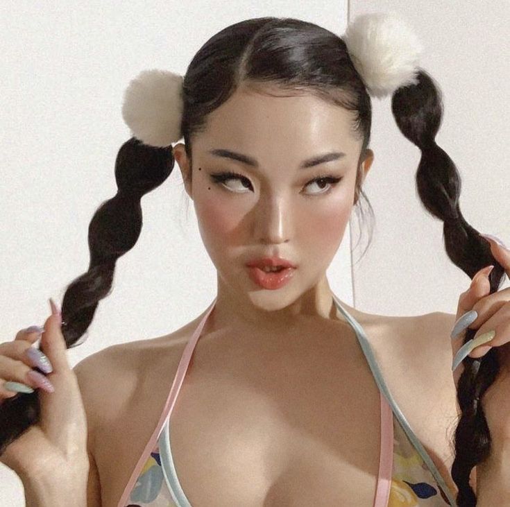 a young asian woman is looking away from the camera while holding a strand of her braided hair. she has her hair in two pigtails, each with a white fluffy bow. she's wearing a light pink bikini top and has her makeup done.