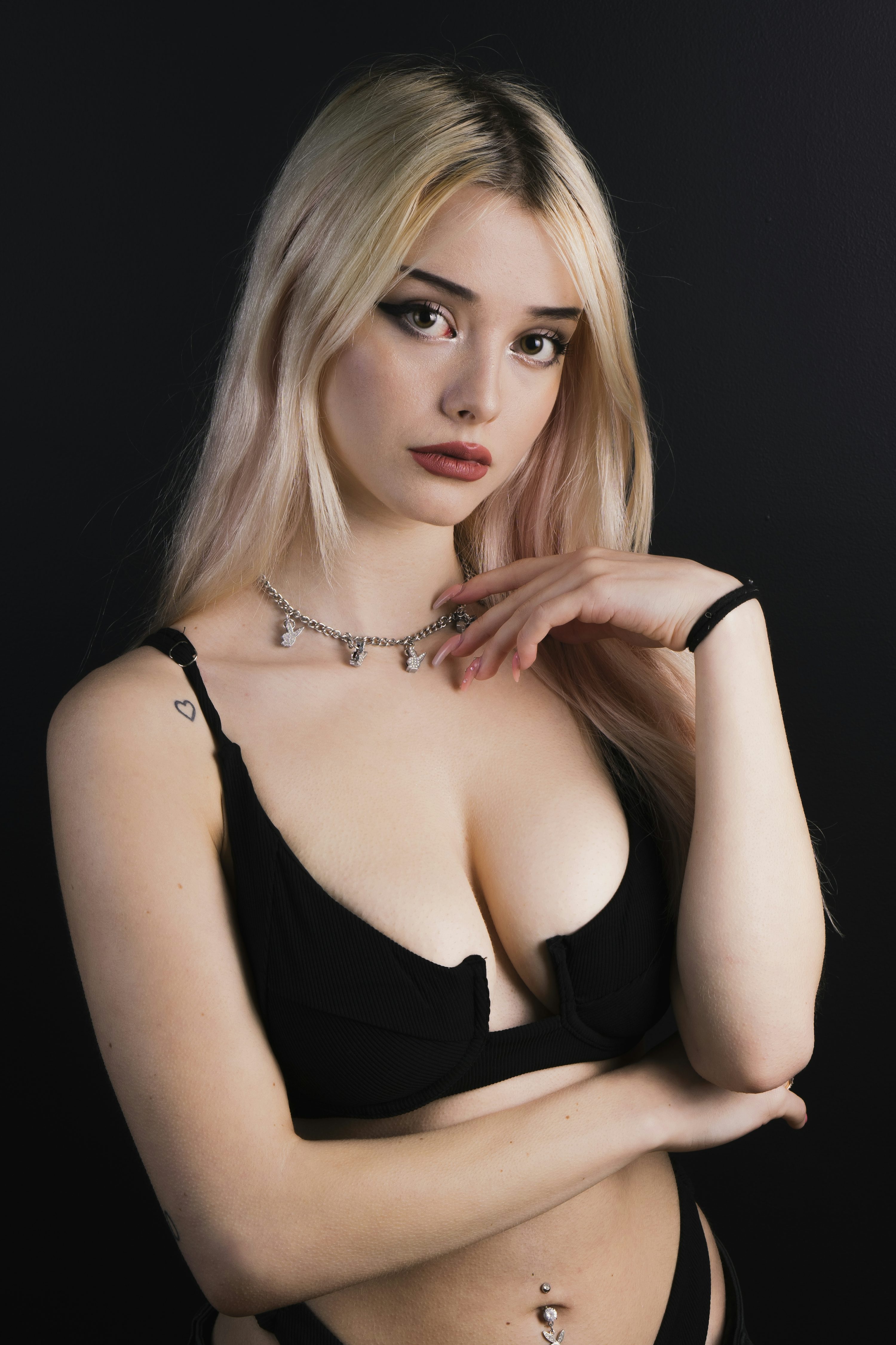 loseup portrait of a young, attractive blonde woman with fair skin. she is wearing a black bra and necklace, and her arms are crossed. her makeup is noticeable, especially the eyeliner and lipstick. the background is black, making her stand out. the overall mood is sensual and beautiful.