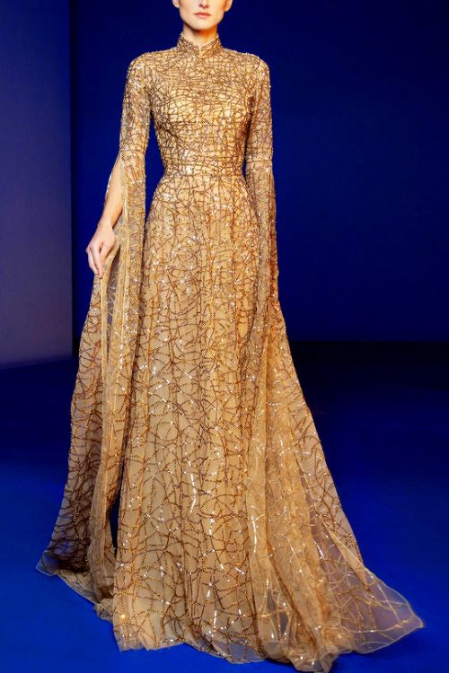a woman is standing and wearing a long, gold, sequined evening gown with long sleeves and a high neck. the gown has a fitted bodice and a flowing skirt. the sleeves have slits. the model is standing against a blue background.