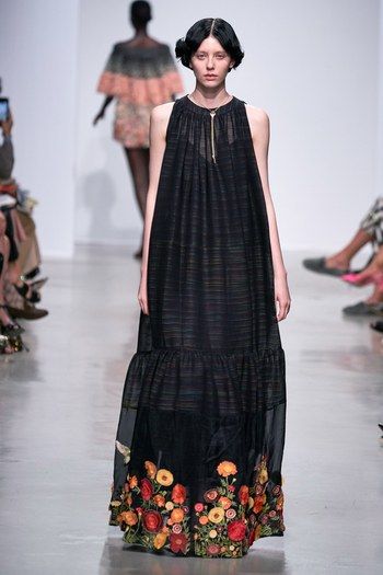 a young woman model in a black, long floral maxi dress with an embroidered floral pattern on the bottom hem, posing on a runway during a fashion show.  she has her dark hair pulled back in a bun and a simple necklace with a gold pendant. the model's outfit is elegant and chic, showcasing a stylish and contemporary look.