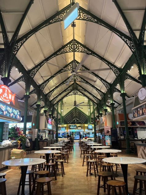 the image shows the interior of a hawker centre, likely in singapore. the architectural style is prominent, featuring a high ceiling with ornate green metalwork.  numerous empty marbletopped tables and wooden stools are arranged in neat rows down the center aisle.  the food stalls are visible along the sides, but most appear unoccupied. the overall atmosphere is calm and quiet, despite the building's potential for bustling activity.  the lighting is bright, and large ceiling fans are visible.
