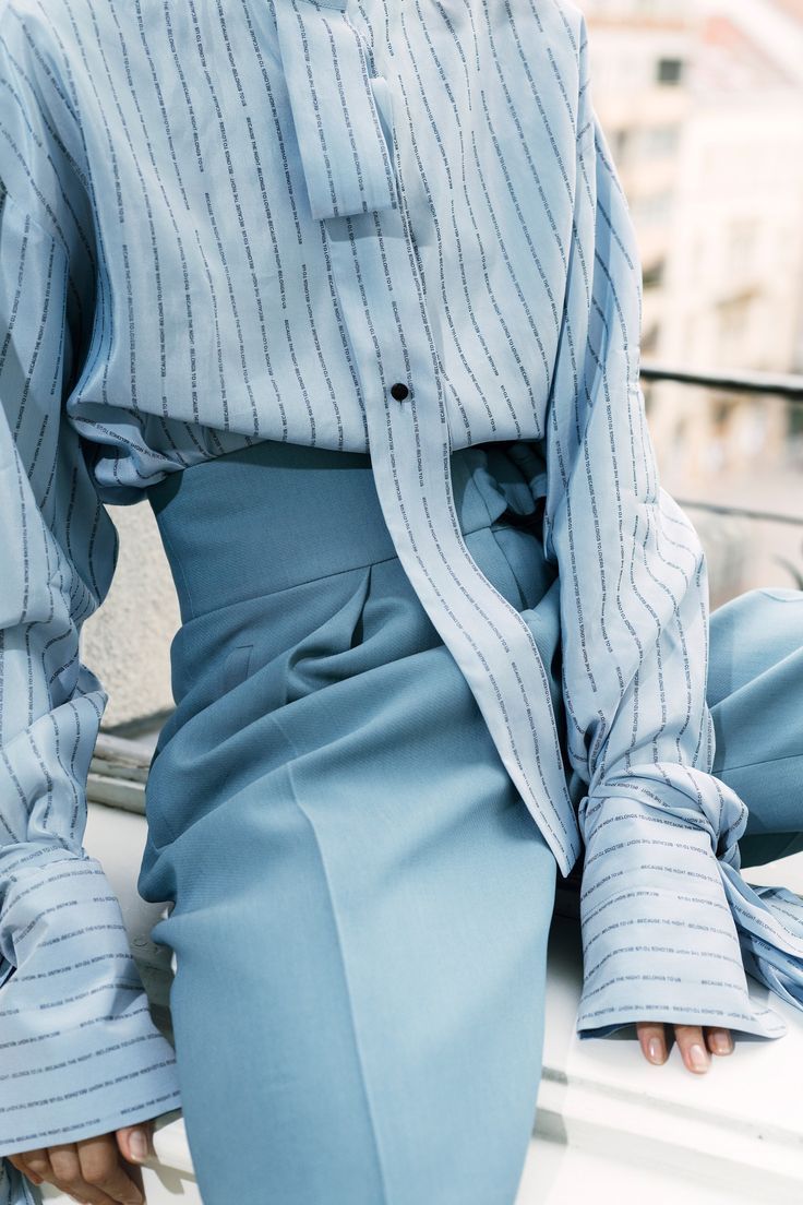 the image is a closeup shot focusing on the details of a woman's outfit. she is wearing a pale blue pinstripe oversized shirt with rolledup sleeves, paired with highwaisted, wideleg trousers in a matching light blue shade. the outfit is monochromatic, creating a minimalist and sophisticated look. the focus is on the texture and drape of the fabric, highlighting the elegant and loose fit of both the shirt and pants. the image showcases a detail shot that emphasizes the style and quality of the clothing. the background is blurred, drawing attention to the outfit itself.