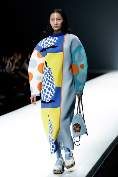 a fashion model walks the runway wearing a striking, colorful dress. the dress features a patchwork design with polka dots, geometric shapes, and a large pear motif. the colors are predominantly blue, yellow, and white, with accents of orange and grey. the model also carries a small, light blue handbag with colorful embellishments.  her shoes have a similar playful, colorful design. the overall aesthetic is modern, artistic, and avantgarde. the background is a simple, clean runway setting.
