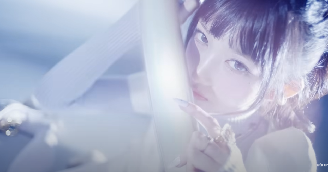 a closeup image of a young woman with dark hair and bangs. she is wearing a white turtleneck sweater and silver rings. her nails are long and painted white. she is lit by a bright, cool light source that highlights her features and creates a soft, ethereal look. the overall color scheme of the image is cooltoned, with shades of blue and white dominating.  the background is blurred, suggesting a shallow depth of field and further emphasizing the subject.