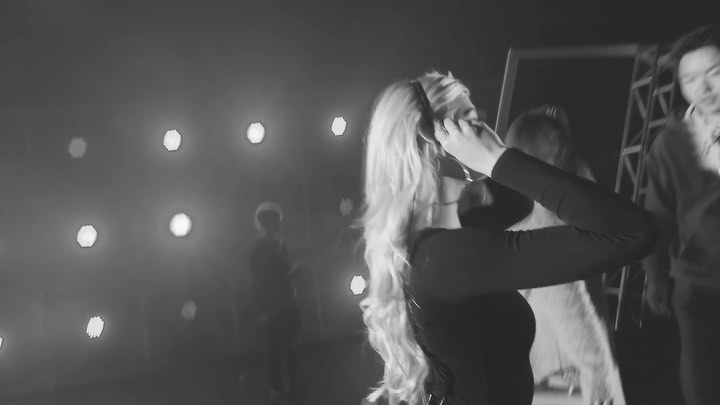 a black and white image shows a woman with long blonde hair putting on headphones. she is wearing a dark, fitted top. behind her are other people and stage equipment. in the background are multiple bright lights arranged in a pattern. the scene appears to be backstage at a performance or event.