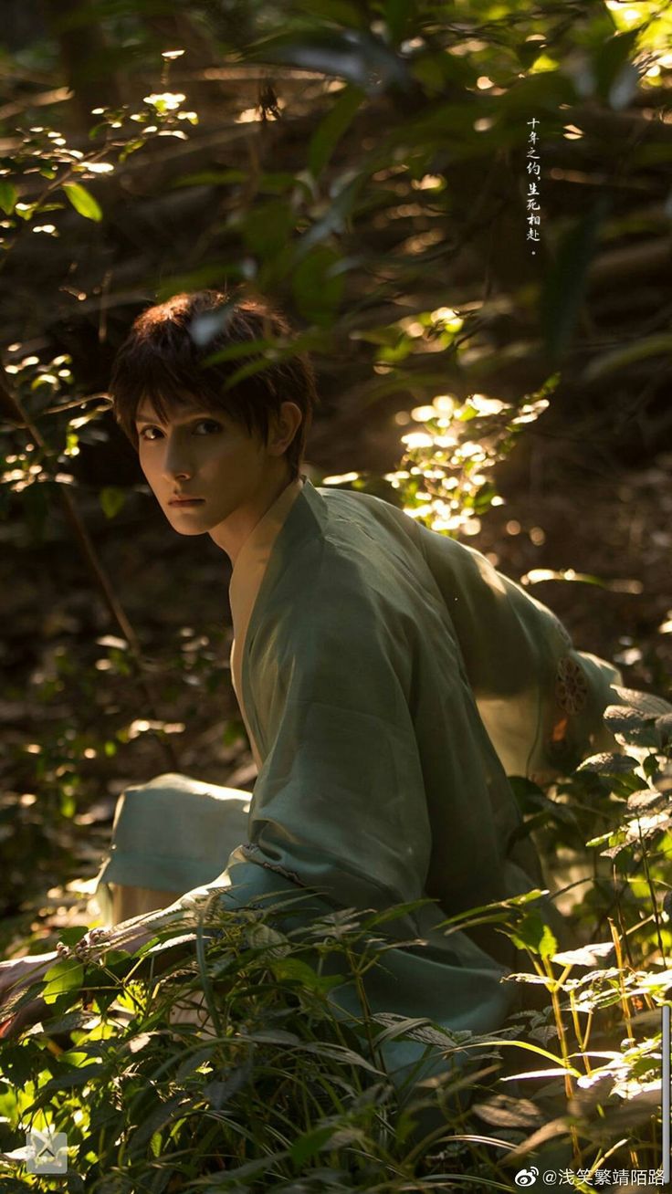 a man in a teal kimono crouches in a forest. sunlight filters through the trees, casting dappled light on him and the surrounding foliage. he is looking back over his shoulder towards the camera. the overall atmosphere is serene and slightly mysterious.