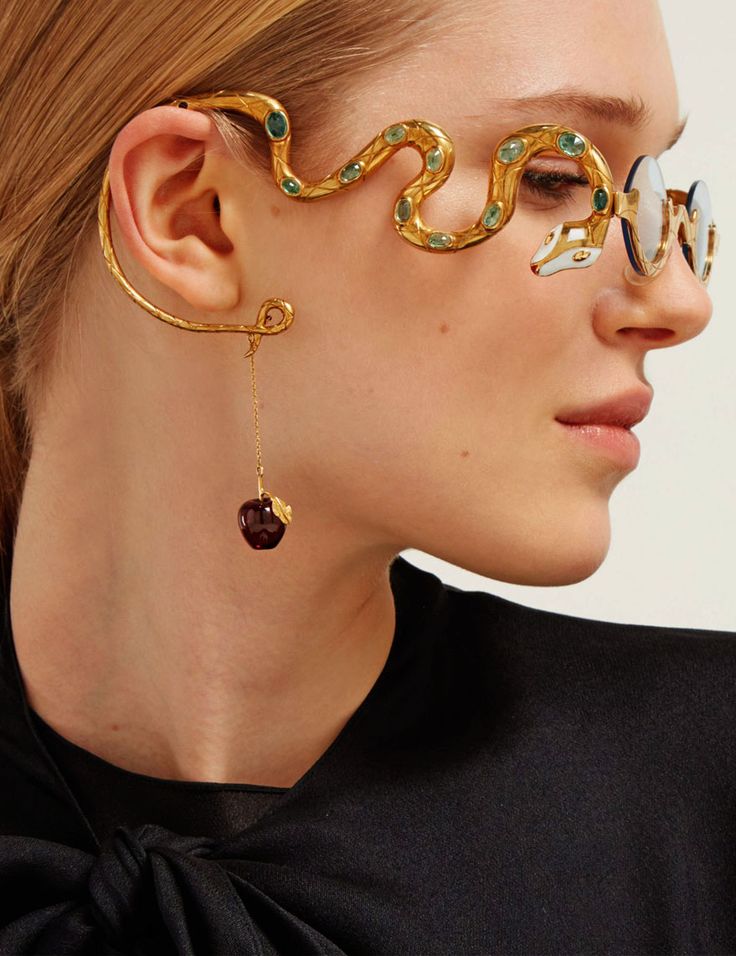 closeup view of a woman's face and neck, showcasing a pair of unique gold snakeshaped glasses and an elaborate gold snake ear cuff with a dangling dark red apple charm.  the woman has long light brown hair and is wearing a black top. the focus is sharp on the jewelry and the model's face. the image highlights the intricate detail and luxurious feel of the accessories.