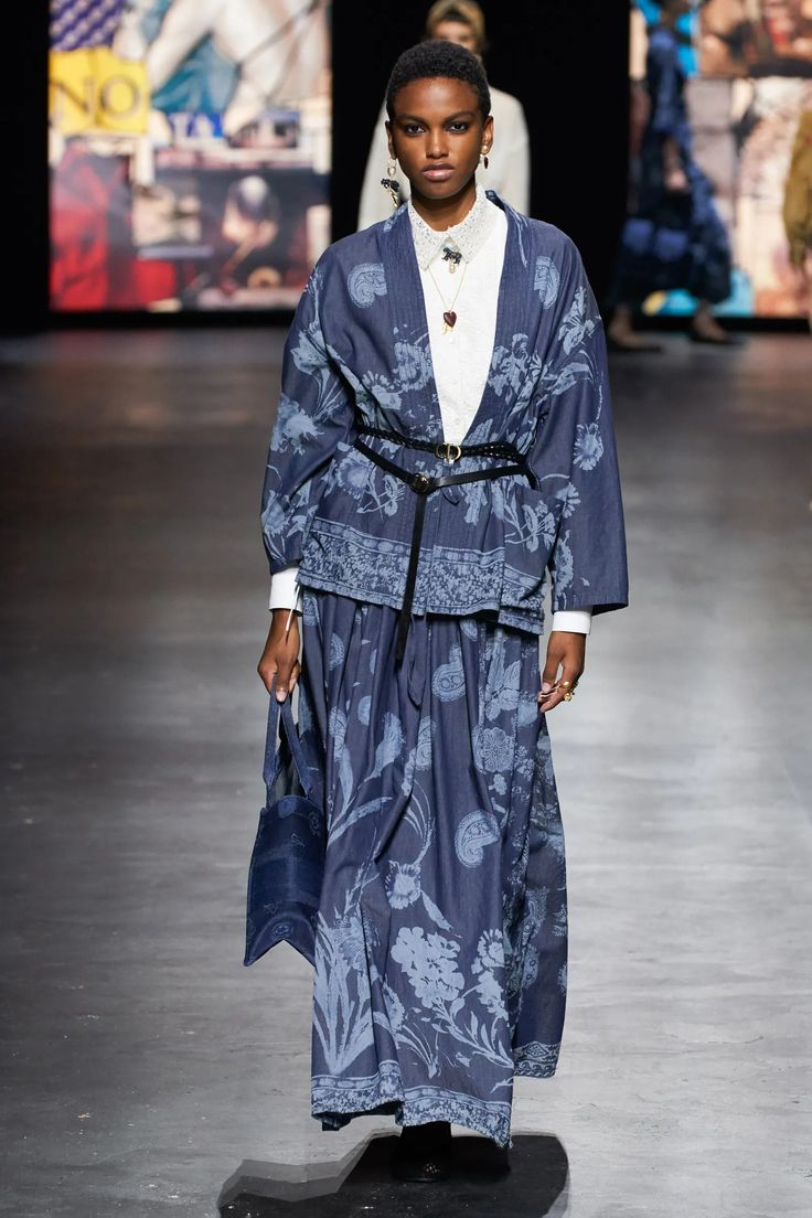 a fashion model walks down the runway wearing a long denim skirt and matching jacket with a floral print. she has a white shirt underneath, a black belt with a gold buckle, and a blue bag. the model is posing for the camera and walking confidently down the runway.