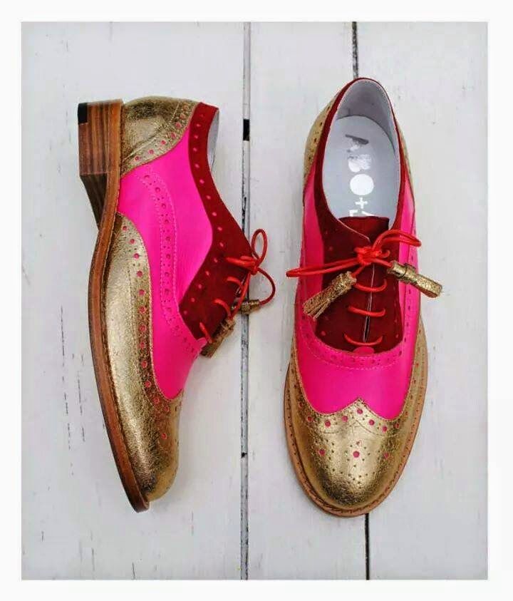 the image shows a pair of women's brightly colored oxfords or brogues. the shoes are predominantly pink and gold, with a fuchsia pink section and metallic gold detailing.  the laces are burgundy with small gold tassels.  the shoes appear to be leather and are styled in a classic wingtip pattern. they are positioned on a lightcolored wooden surface. one shoe is positioned at a slight angle while the other is laid flat, displaying the full design of the footwear. the brand name "bo+bo" might be faintly visible on the inside of one shoe.